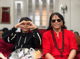 Young girl and an older woman sit on a red couch; the girl forms a heart with her hands around her face as they smile at the camera.