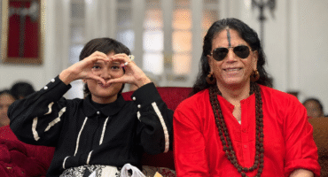 Young girl and an older woman sit on a red couch; the girl forms a heart with her hands around her face as they smile at the camera.