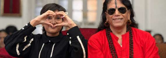 Young girl and an older woman sit on a red couch; the girl forms a heart with her hands around her face as they smile at the camera.